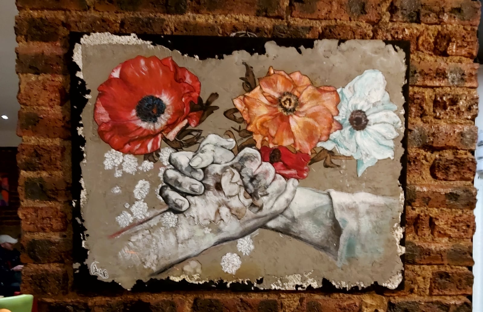 Tableau accroché sur mur de briques — mains tenant trois fleurs